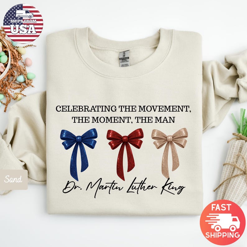 Peut inclure: Sweat-shirt cr&egrave;me avec le texte "CELEBRATING THE MOVEMENT, THE MOMENT, THE MAN" et "Dr. Martin Luther King." En dessous, trois n&oelig;uds bleus, rouges et beiges. Le sweat-shirt a un col rond.