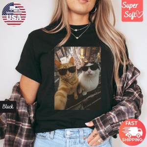 May include: Black t-shirt featuring a selfie of two cats wearing sunglasses. One cat is orange, the other is white and black. The shirt has a "Super Soft" label and a "Fast Shipping" label.