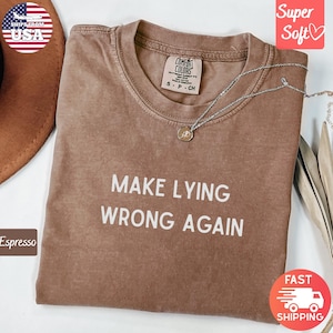 Może przedstawiać: T-shirt w kolorze espresso z białym napisem "MAKE LYING WRONG AGAIN". Koszulka ma okrągły dekolt i krótkie rękawy. Srebrny naszyjnik z wisiorkiem jest zarzucony na koszulkę. Koszulka jest oznaczona jako "Super Soft".