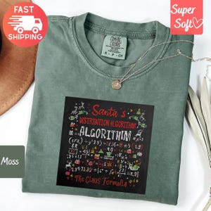 May include: A moss green Comfort Colors t-shirt with a black graphic featuring the text "Santa's Distribution Algorithm" and mathematical equations. The shirt has a crew neck and short sleeves. A silver necklace with a pendant is draped on the shirt.