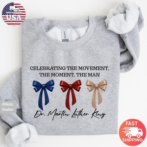 Peut inclure: Sweat-shirt gris chin&eacute; avec l'inscription "CELEBRATING THE MOVEMENT, THE MOMENT, THE MAN" et "Dr. Martin Luther King". Il pr&eacute;sente trois n&oelig;uds bleus, rouges et dor&eacute;s.