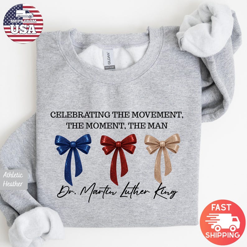 Peut inclure: Sweat-shirt gris chin&eacute; avec l'inscription "CELEBRATING THE MOVEMENT, THE MOMENT, THE MAN" et "Dr. Martin Luther King". Il pr&eacute;sente trois n&oelig;uds bleus, rouges et dor&eacute;s.