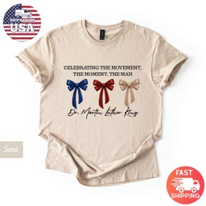 Peut inclure: T-shirt couleur sable avec l'inscription "CELEBRATING THE MOVEMENT, THE MOMENT, THE MAN" et "Dr. Martin Luther King." Le motif comprend trois n&oelig;uds papillon bleus, rouges et beiges. Le t-shirt a un col rond et des manches courtes.
