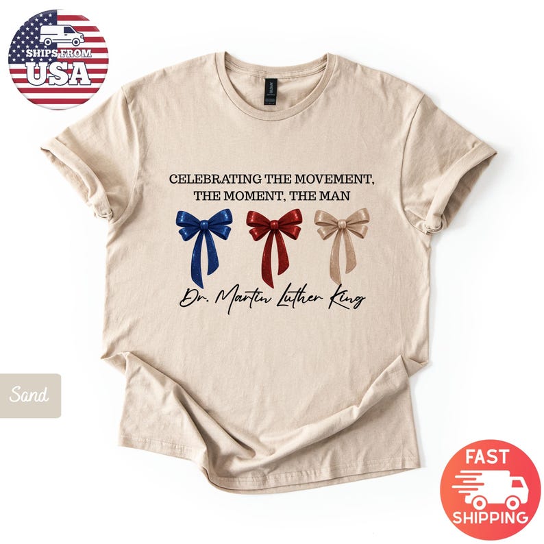Peut inclure: T-shirt couleur sable avec l'inscription "CELEBRATING THE MOVEMENT, THE MOMENT, THE MAN" et "Dr. Martin Luther King." Le motif comprend trois n&oelig;uds papillon bleus, rouges et beiges. Le t-shirt a un col rond et des manches courtes.