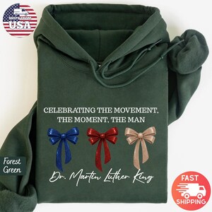 Peut inclure: Sweat &agrave; capuche vert for&ecirc;t avec le texte "CELEBRATING THE MOVEMENT, THE MOMENT, THE MAN" et "Dr. Martin Luther King". Comprend trois motifs de n&oelig;uds bleus, rouges et beiges. Un sweat &agrave; capuche.