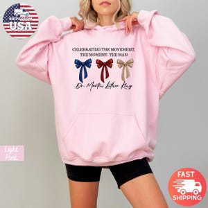 Peut inclure: Sweat &agrave; capuche rose clair avec une poche avant et une capuche &agrave; cordon. Le devant pr&eacute;sente trois graphiques de n&oelig;uds papillon bleus, rouges et beiges, avec le texte "CELEBRATING THE MOVEMENT, THE MOMENT, THE MAN Dr. Martin Luther King."