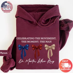 Peut inclure: Sweat &agrave; capuche bordeaux avec le texte "CELEBRATING THE MOVEMENT, THE MOMENT, THE MAN" et "Dr. Martin Luther King." Comporte trois n&oelig;uds d&eacute;coratifs bleus, rouges et dor&eacute;s. Le sweat &agrave; capuche est pli&eacute;.