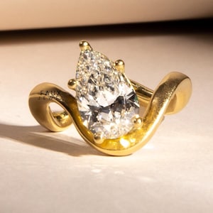Peut inclure: Une bague en or avec un diamant en forme de poire. La bague a un design unique et fluide avec le texte "FAUX JEWEL" visible sur le côté. Le diamant est clair et scintillant, serti dans une monture en or.