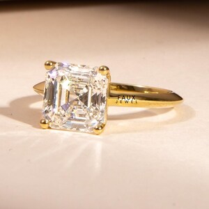 Puede incluir: Un anillo de oro con una gran piedra preciosa clara de forma rectangular. La piedra est&aacute; engastada con cuatro peque&ntilde;as garras de oro. La banda del anillo es de oro pulido simple. El texto "FAUX JEWEL" es visible en el anillo.