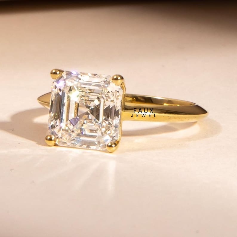 Puede incluir: Un anillo de oro con una gran piedra preciosa clara de forma rectangular. La piedra est&aacute; engastada con cuatro peque&ntilde;as garras de oro. La banda del anillo es de oro pulido simple. El texto "FAUX JEWEL" es visible en el anillo.