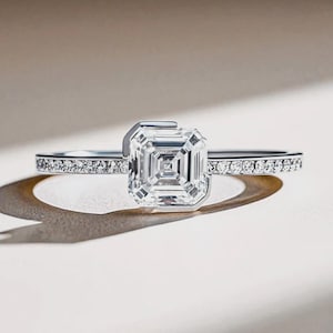 May include: A silver ring featuring a large, square-cut, clear gemstone. The band is embellished with smaller, round gemstones. The ring is set against a neutral backdrop, with the text "FAUX JEWEL" visible on the band.