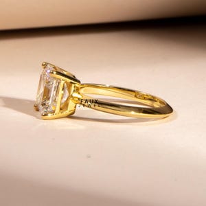 Puede incluir: Un anillo de oro con una gema grande, rectangular y transparente. La banda y el engaste del anillo son de color dorado brillante. Las palabras "FAUX JEWEL" son visibles en el anillo.