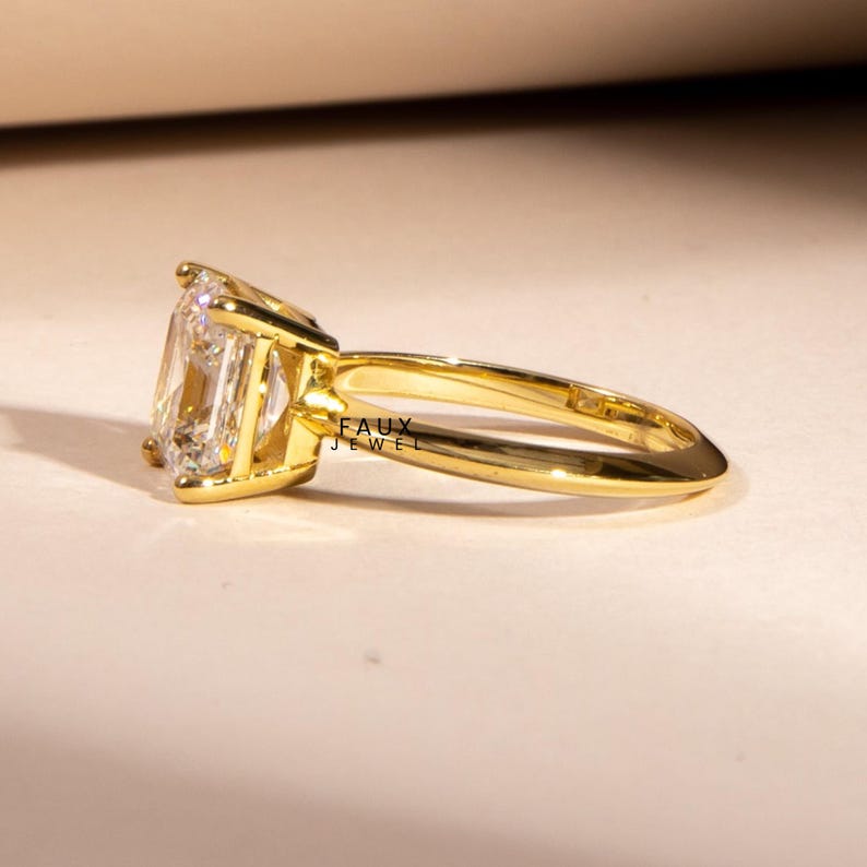 Puede incluir: Un anillo de oro con una gema grande, rectangular y transparente. La banda y el engaste del anillo son de color dorado brillante. Las palabras "FAUX JEWEL" son visibles en el anillo.