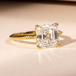 Puede incluir: Un anillo de oro con una gran gema transparente de corte cuadrado. La banda del anillo es fina y pulida, con el texto "FAUX JEWEL" grabado en el lateral. La gema est&aacute; sujeta por cuatro garras de oro.