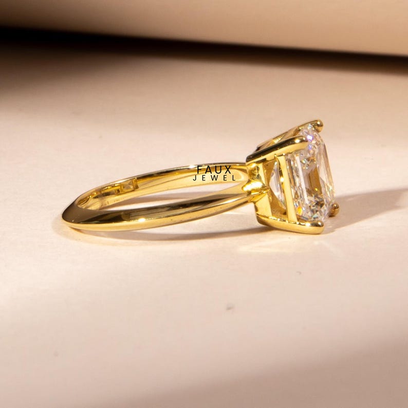 Puede incluir: Un anillo de oro con una gran gema clara de forma rectangular. La banda del anillo es delgada y pulida, con el texto "FAUX JEWEL" grabado en el lateral. La gema est&aacute; engastada en un bisel de oro.