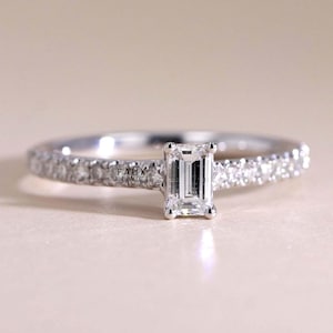 May include: A silver ring with a rectangular, emerald-cut diamond center stone. The band is encrusted with small, round diamonds. The ring is set against a soft, neutral background. The text "FAUX JEWEL" is visible on the side of the ring.