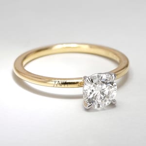 May include: A gold band engagement ring with a large, round, clear gemstone. The gemstone is held in place by four silver prongs. The ring is set against a white background. The text "FAUX JEWEL" is printed on the band.