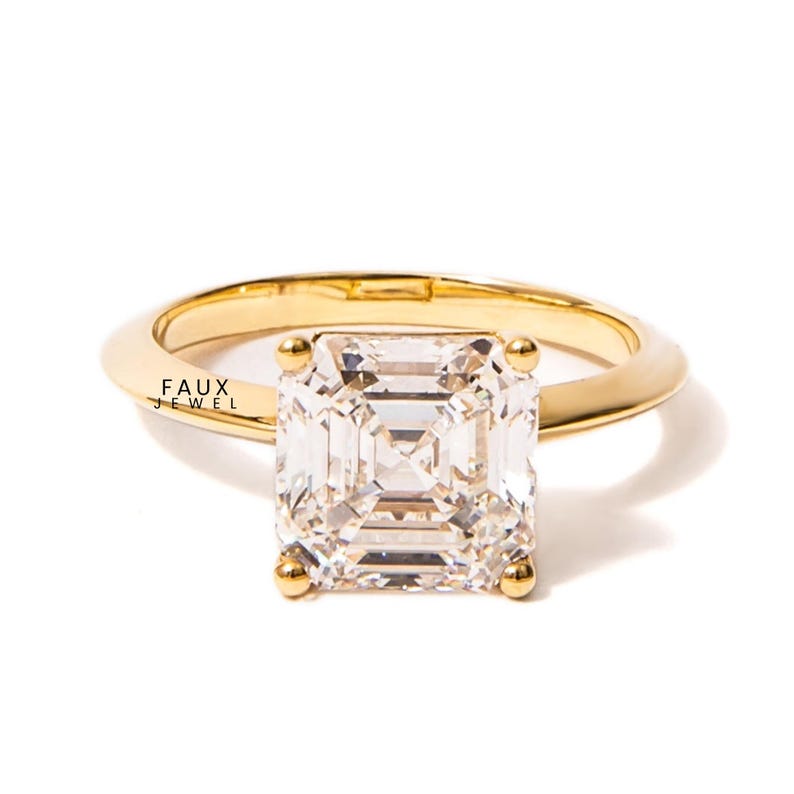 Puede incluir: Un anillo de oro con una gran gema transparente de corte cuadrado. La banda del anillo es lisa y redondeada. La gema est&aacute; sujeta por cuatro peque&ntilde;as garras doradas. El texto "FAUX JEWEL" es visible.