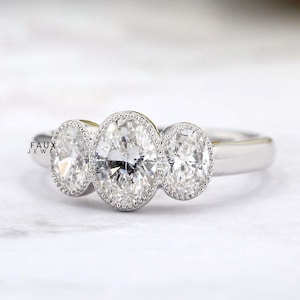 May include: A three-stone engagement ring with oval-cut diamonds. The ring features a delicate band and each diamond is surrounded by a halo of smaller stones. The ring is set against a white background.
