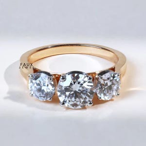 May include: A gold-toned ring featuring three large, clear, round-cut stones. The stones are set in a classic prong setting, and the band is smooth and polished. The ring is displayed on a white surface.