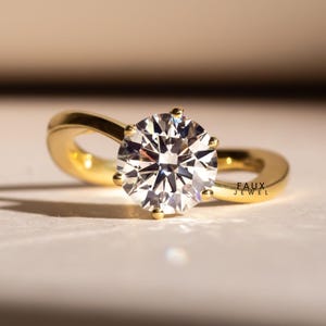 May include: A gold engagement ring featuring a large, round, clear gemstone. The ring has a simple, elegant design with a swirling band. The text "FAUX JEWEL" is visible on the ring.