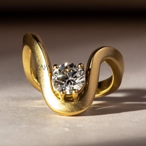 May include: A gold ring with a clear, round gemstone. The ring has a distinctive, flowing design that curves around the central stone. The text "FAUX JEWEL" is visible on the ring.