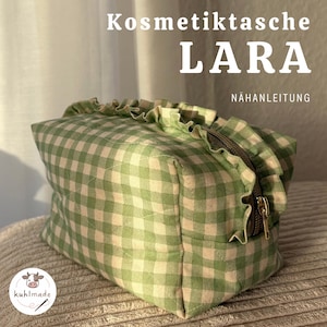 May include: A green and white gingham cosmetic bag with a ruffled top and a gold zipper. The bag is labeled "Kosmetiktasche LARA" and "NÄHANLEITUNG". The bag is sitting on a textured surface.