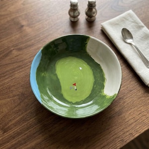 Può includere: Una ciotola in ceramica con un design da campo da golf. La ciotola presenta un fairway verde, un cielo blu e una sabbia bianca. Una piccola bandiera rossa e bianca segna la buca. La ciotola è su un tavolo di legno.
