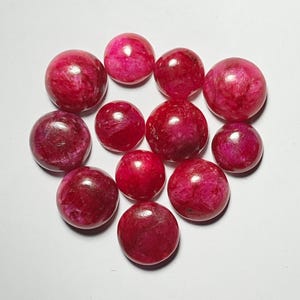 May include: A collection of eleven polished, round gemstones in varying shades of red. The stones have a smooth, reflective surface, with a marbled appearance. The gemstones are arranged on a white surface, showcasing their color and texture.