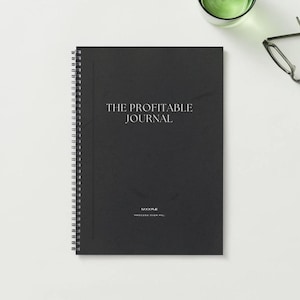 Könnte beinhalten: Ein schwarzes Spiralbuch mit dem Titel "THE PROFITABLE JOURNAL" in weißer Schrift. Das Notizbuch liegt auf einer weißen Oberfläche, mit einer Brille und einem Glas grüner Flüssigkeit im Hintergrund.