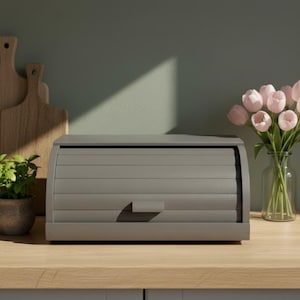 May include: A grey bread bin with a roll-top lid on a wooden surface. A small potted plant and two wooden chopping boards are to the left. A vase of pink tulips is to the right. The background is a muted green.