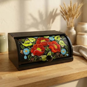 Puede incluir: Una panera de madera negra con tapa enrollable que presenta un diseño floral pintado a mano. La obra muestra amapolas rojas vibrantes, pequeñas flores azules y hojas verdes sobre un fondo oscuro, con un pomo negro para su apertura.