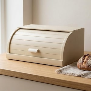 May include: A cream-colored wooden bread box with a roll-top lid and a small handle. The box sits on a wooden countertop next to a loaf of bread on a linen cloth. The bread box has a scalloped edge.