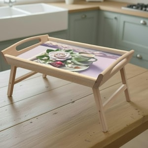 Puede incluir: Una bandeja de desayuno de madera clara con un diseño floral. La bandeja tiene una parte superior rectangular con una imagen impresa de una rosa, una taza de té y un platillo. La bandeja está sostenida por patas plegables.