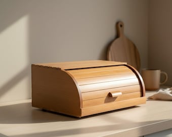 May include: A rectangular wooden bread box featuring a curved, slatted roll-top lid with a small handle. The light-toned wood grain is visible across the horizontal slats of the lid and the flat side panels of this kitchen storage container.