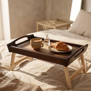 May include: A dark brown wooden breakfast tray with light-colored legs holds a cup of coffee, a croissant on a plate, and a small vase of flowers. The tray is set on a bed with neutral-toned bedding.