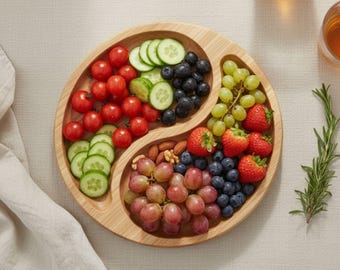 May include: A round bamboo serving tray shaped like a yin-yang symbol, divided into two sections. One side holds red cherry tomatoes and sliced cucumbers, while the other contains green grapes, red strawberries, blueberries, and almonds.