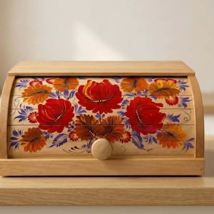 Peut inclure: Une boîte à pain en bois clair avec une porte coulissante ornée d'un motif floral peint à la main. L'illustration présente de grandes fleurs rouges et orange avec des feuilles vertes et bleues. Un petit bouton rond en bois est centré.