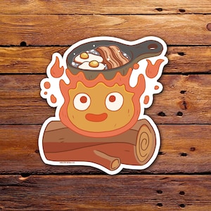 Puede incluir: Una pegatina con un personaje de dibujos animados hecho de fuego, con una cara sonriente y llamas. Una sartén con huevos y tocino se encuentra encima de las llamas. Fondo de madera.