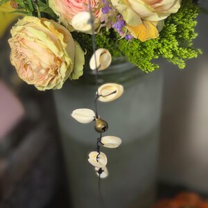 May include: A close-up of a floral arrangement with yellow and pink roses, green foliage, and small purple flowers. A string of white seashells with a small brass bell hangs in front of the flowers.