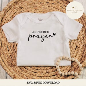 May include: White baby onesie with the words "ANSWERED prayer" in black script, a black heart, and the Gerber brand label. The onesie is on a woven wicker surface with a string of wooden beads.