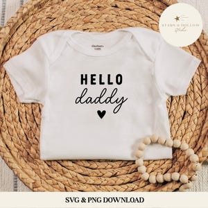 May include: White baby bodysuit by Gerber, featuring the text "HELLO daddy" in black script, a black heart, and the brand logo. The bodysuit is displayed on a woven wicker surface.