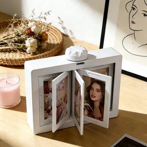 Puede incluir: Un marco de fotos blanco con un botón circular en la parte superior. El marco muestra varias fotos, incluyendo un retrato de mujer y un oso de peluche. También se ven una vela rosa encendida y una cesta de mimbre con flores secas.