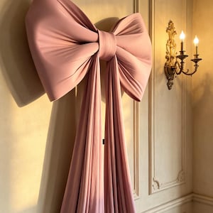 Puede incluir: Un gran lazo de tela rosa empolvado montado en una pared. El lazo presenta dos bucles anchos y estructurados en la parte superior y dos largas colas de tela fluida que caen verticalmente, creando pliegues suaves y una textura mate.