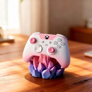 May include: A white and pink Xbox controller with pink buttons and a black Xbox logo, resting on a decorative stand of blue, pink, and purple crystal-like formations. The controller is on a wooden surface.