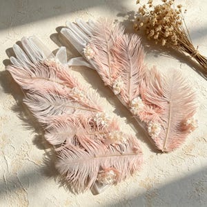 May include: A pair of sheer white gloves adorned with long, soft pink feathers and small clusters of white floral embellishments with pearl centers. The feathers are arranged along the length of the gloves, creating a layered, textured appearance.