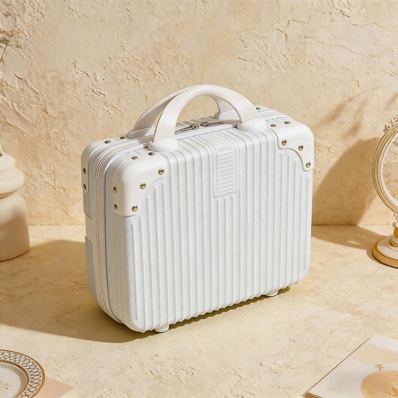 May include: A white, small suitcase with a top handle and gold-colored corner accents. The suitcase has vertical ribbed detailing and a white handle. The overall design is clean and modern, suitable for travel or storage.