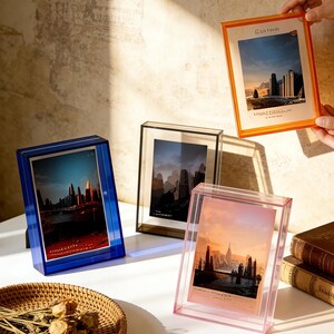 May include: A selection of acrylic picture frames in blue, pink, black, and orange. Each frame contains a landscape print. The frames are displayed on a white surface with a woven basket and books.