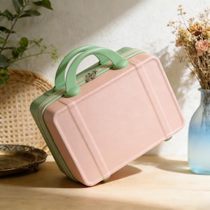 May include: A small, pink suitcase with a mint green handle and trim. The suitcase has a rectangular shape with vertical lines on the front and a silver zipper. The handle is arched, and the suitcase appears to be made of a hard material.