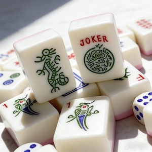 Peut inclure: Un ensemble de tuiles de Mahjong blanches avec des bords roses. Certaines tuiles pr&eacute;sentent des illustrations de dragons et d'oiseaux verts, tandis que d'autres affichent le mot "JOKER" en rouge. Les pi&egrave;ces de jeu sont &eacute;parpill&eacute;es, avec quelques d&eacute;s visibles.